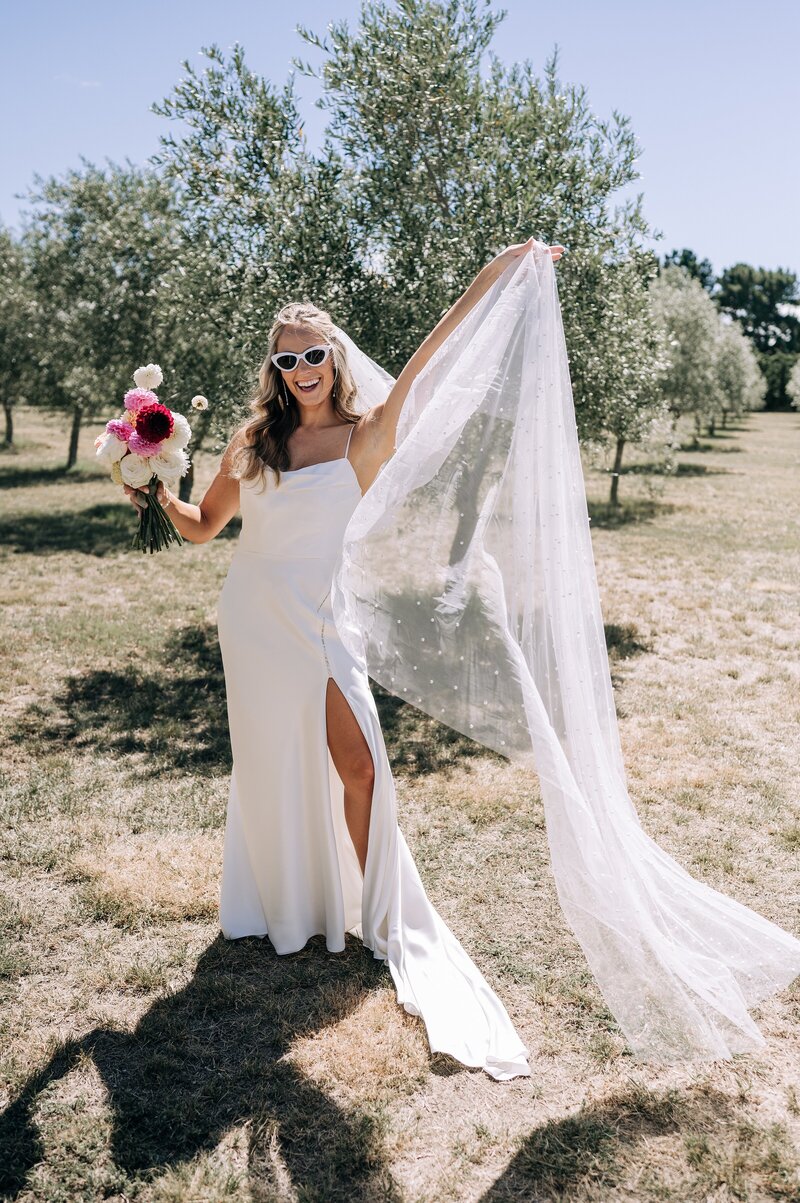 a bride in white sunglasses holds out her pearl studded veil on her wedding day near christchurch at the boneline