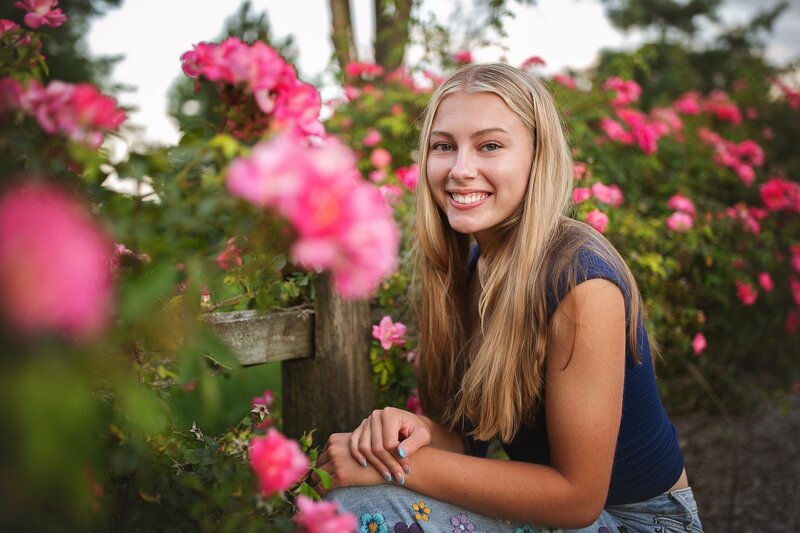 A-smiling-high-school-senior-girl-with-long-blonde-hair-surrounded-by-pink-roses-during-a-summer-photo-session-in-indianapolis-taken-at-flower-farm-Perfect-for-business-professionals-entrepreneurs-and-seniors-celebrating-milestones