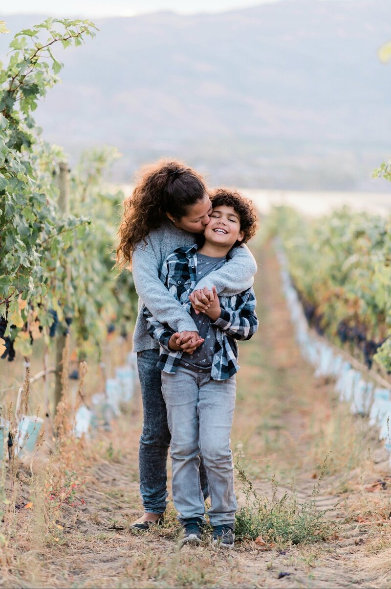 a mom hugging her son, wearing jeans and gray plaid shirt in a vineyard