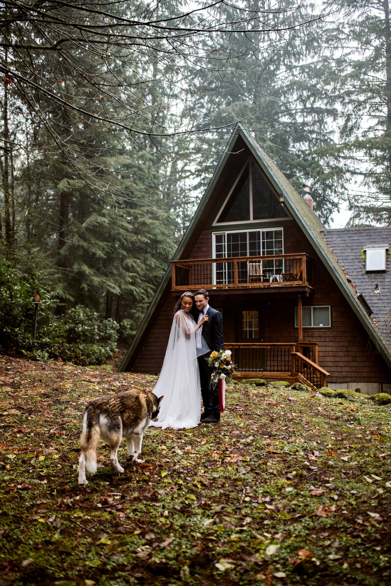 A bride and groom stand outside an A-frame cabin in the Oregon forest with their dog, captured by an Oregon elopement photographer.