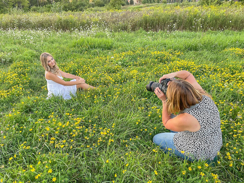 female photographer is sitting on the ground taking a picture of a high school senior girl.  The girl is wearing a white dress and is seated in a green field with yellow flowers