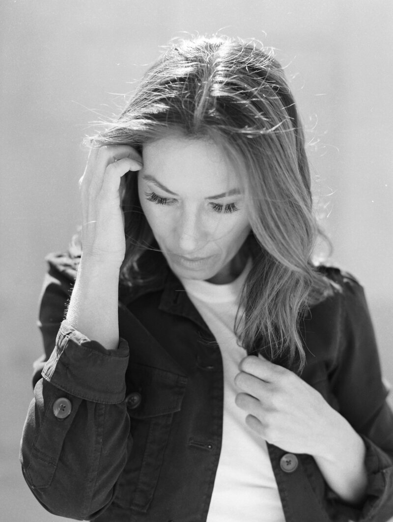 Black and white photo of Michel, she has straight brown hair,a black jacket, and white shirt. She is looking down and adjusting her hair