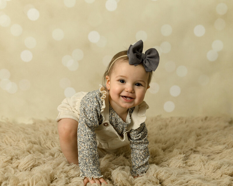 Toddler girl with cream dress. floral shirt and big navy blue bow headband. She's about to stand up and have cream bokeh background behind her.