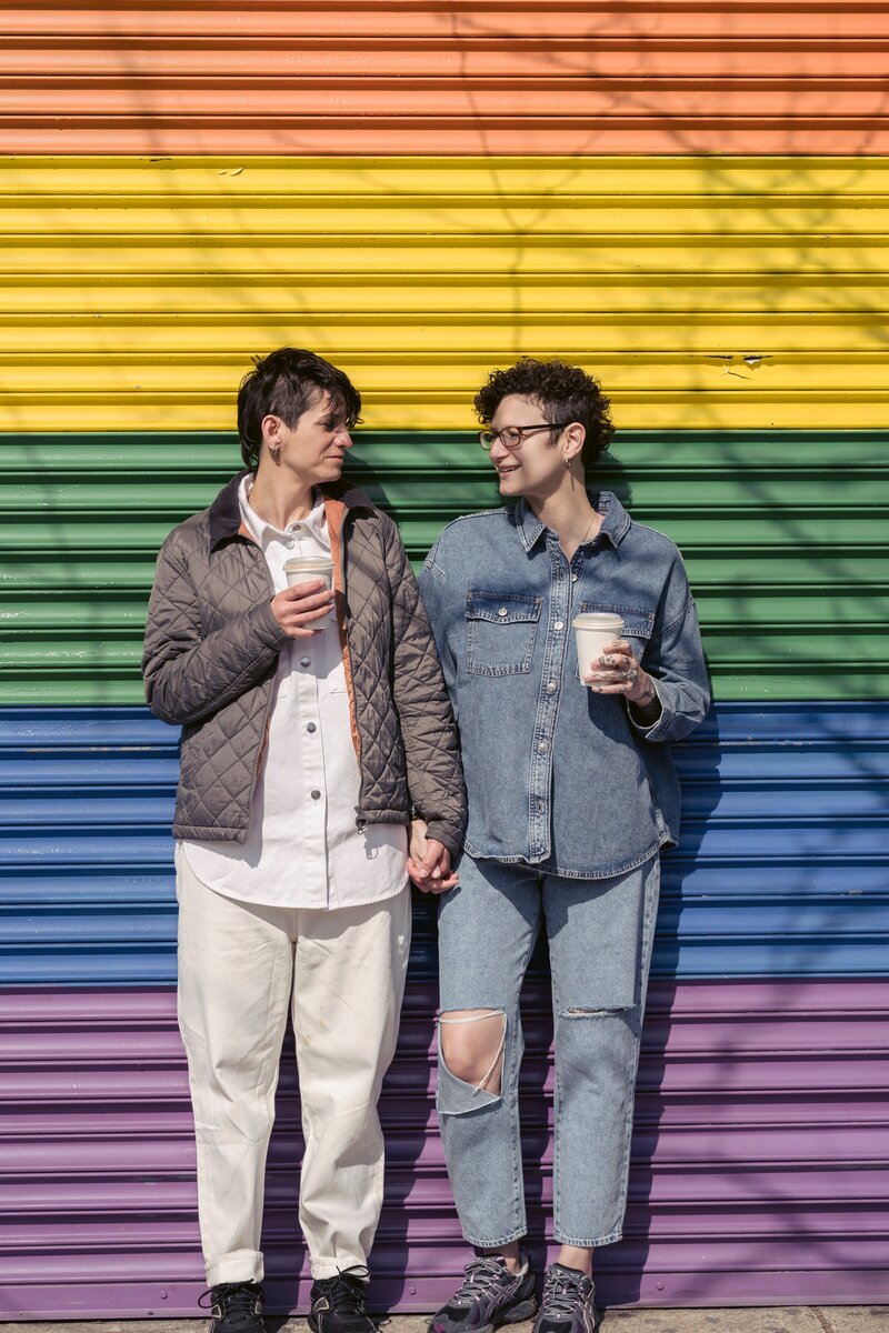 Two feminine presenting partners stand against a rainbow-colored wall, holding hands, with travel coffee cups in their other hands. They face toward each other, smiling.