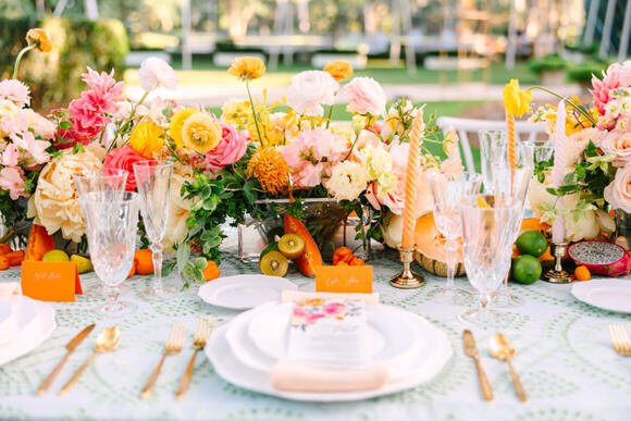This image showcases an elegantly decorated wedding table, featuring vibrant floral arrangements, crystal glassware, and gold flatware. The colorful flowers and fresh fruits create a visually stunning centerpiece, while the delicate lace tablecloth adds a touch of sophistication. Captured by North Carolina wedding photographer Lelia Angelique, this photo highlights the beauty and attention to detail in wedding decor.