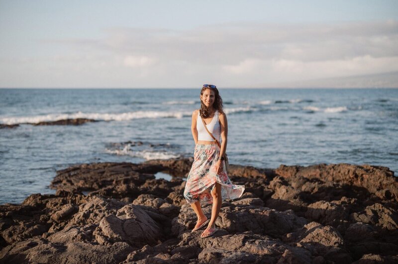Woman stands on black rocks with the ocean behind her