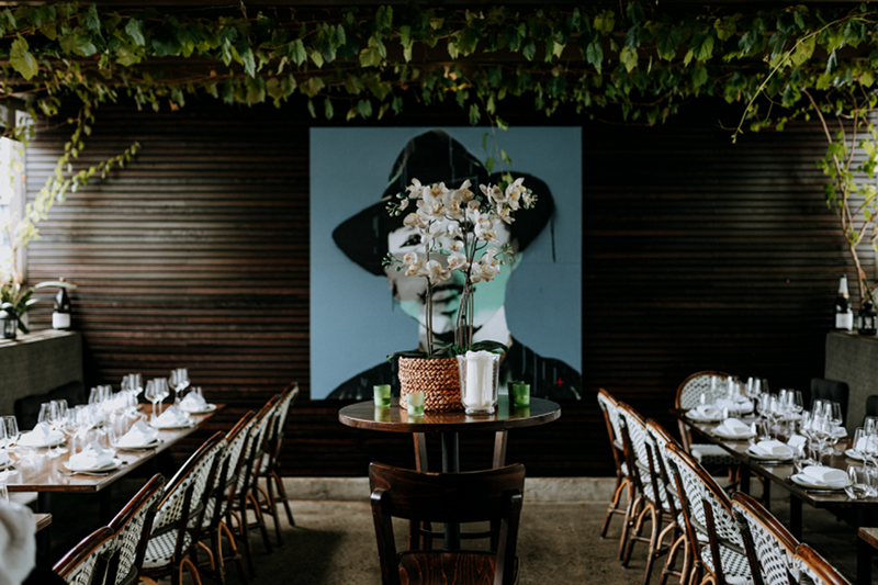 A wine bar with a contemporary artwork of a face hanging behind a set of tables with flowers