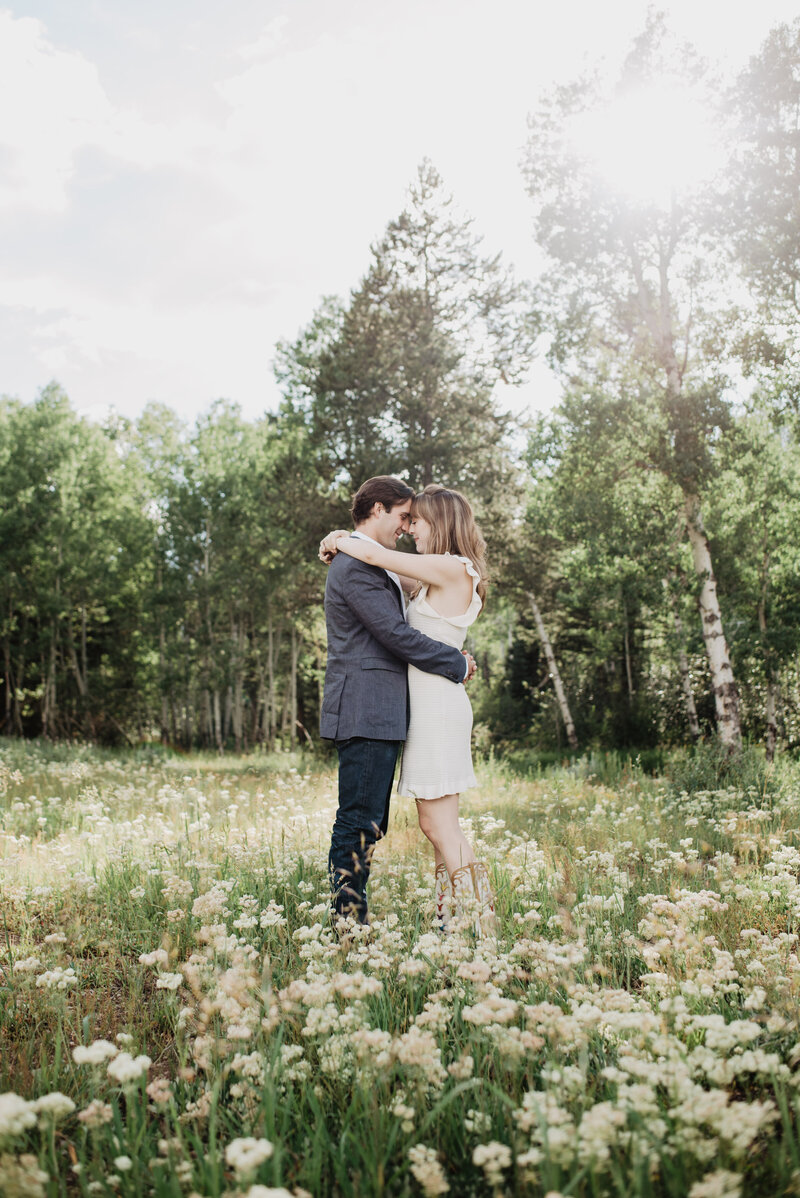 grand teton engagement pictures with man and woman embracing as the fun shines through the tall trees behind them with flowers surrounding them in a meadow