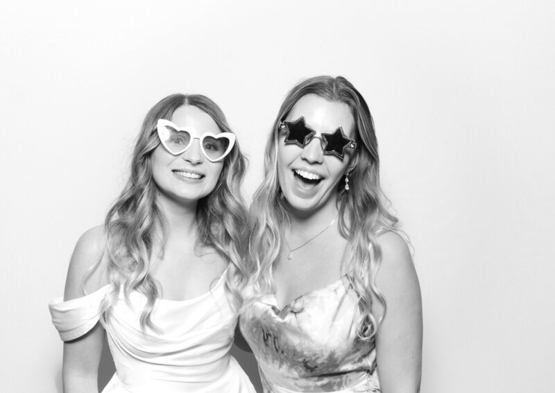 bride and friend during a wedding photo booth experience posing against a white backdrop for a very fun wedding reception entertainment