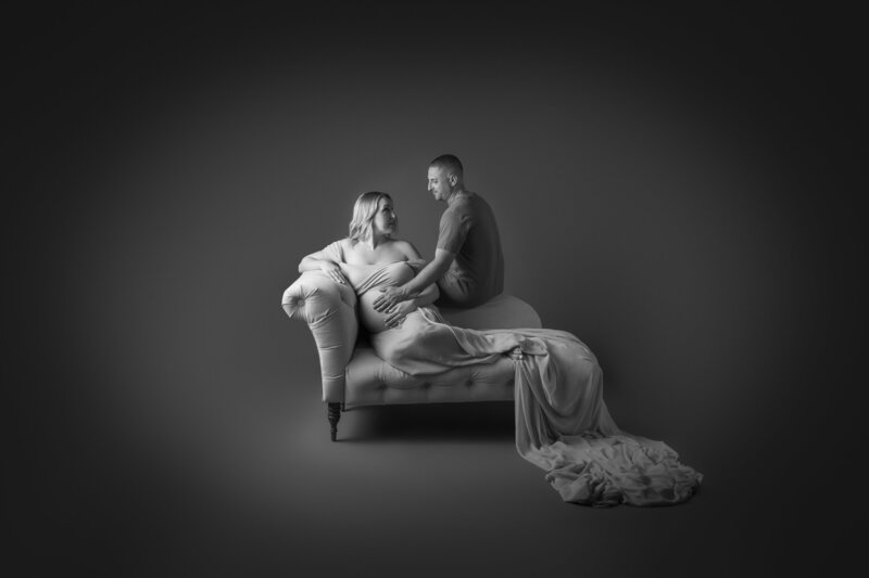 intimate maternity couple photo shoot created in our Fort Mill SC studio, Mom to be in lounging on a fancy chaise lounge and dad to be in perched on the side hand over his wife's belly. Image is black and white. Couple are caucasian
