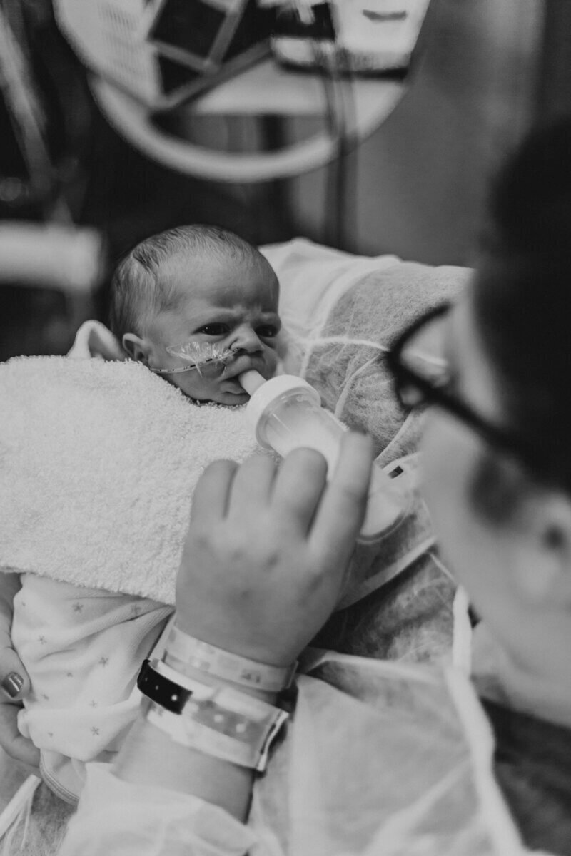 A black and white photo of a newborn being bottle-fed in a hospital, with medical tubes visible and a parent's arm supporting them.