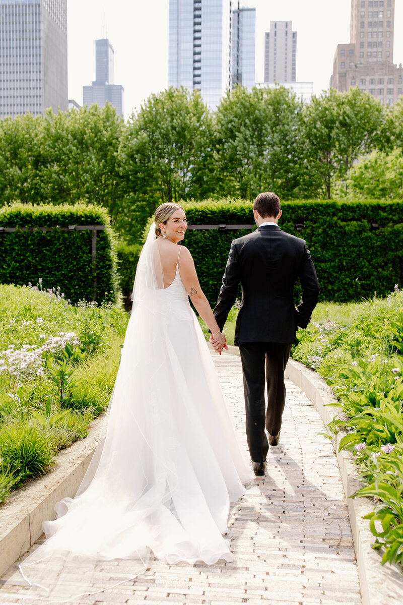 Groom guides his bride down a Chicago park path as she looks back over her shoulder