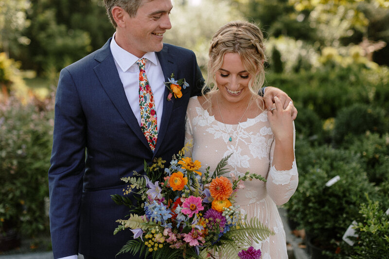 A couple stands together, smiling, with the woman holding a vibrant bouquet of flowers in a lush outdoor setting.