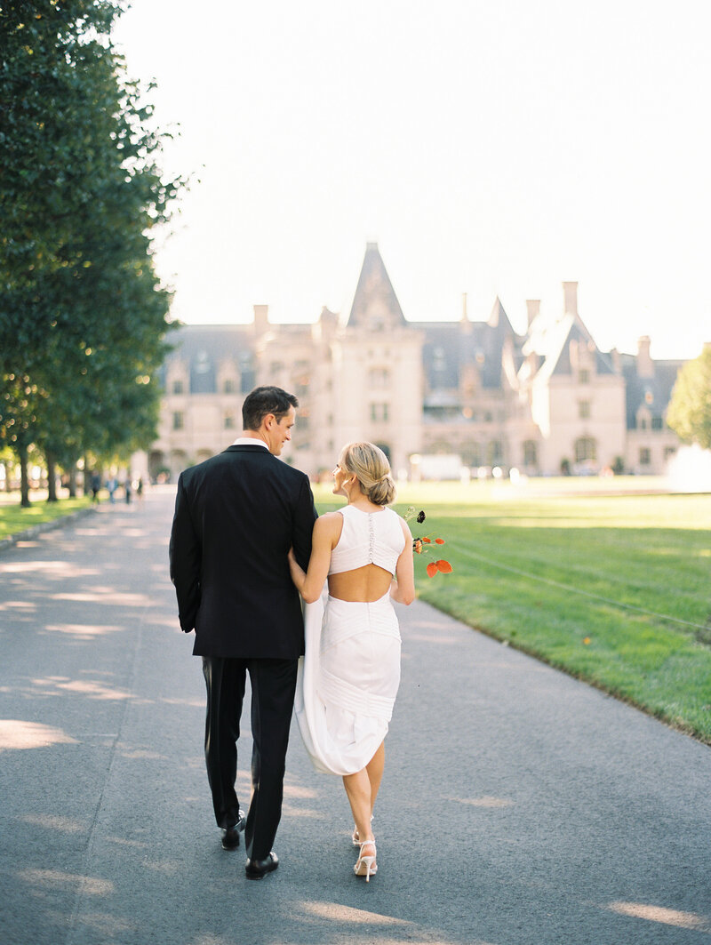 A bride and groom walk towards the Biltmore Estate with their backs to an Asheville Wedding Photographer