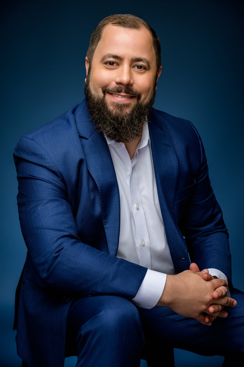 Professional headshot of a man wearing a blue suit, smiling confidently. The image captures a polished and approachable look, ideal for personal branding or business purposes.