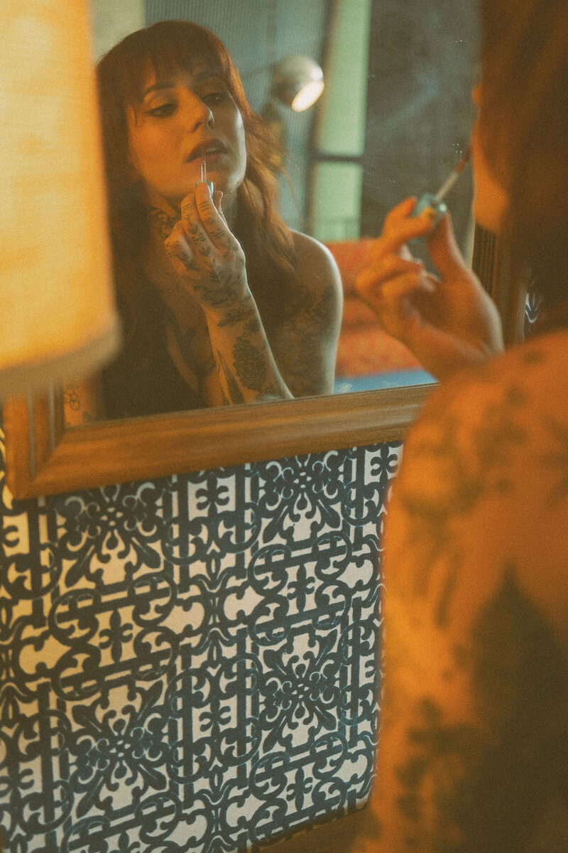A woman with tattoos and a cool vibe putting on lipstick in the mirror for her creative branding photos.