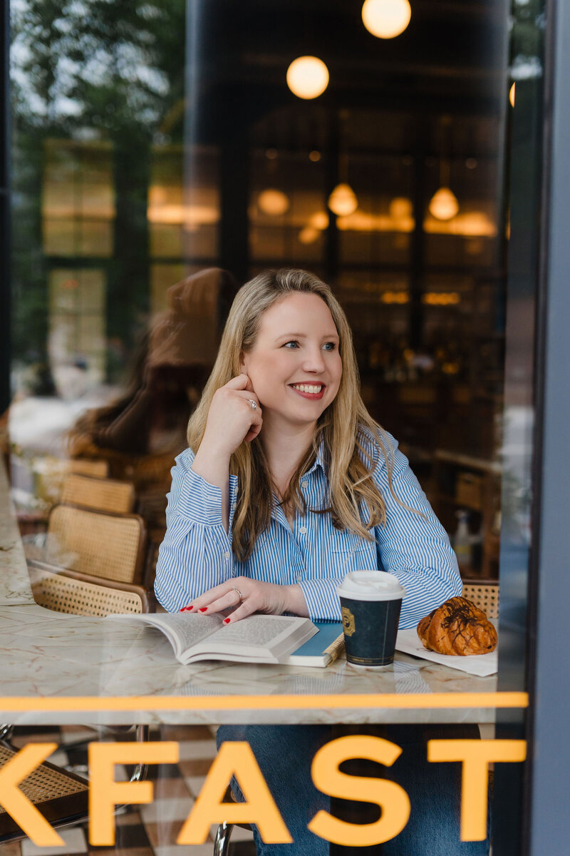 Photo taken of Anna Bradshaw through a cafe window while she sits with a book, coffee, and pastry