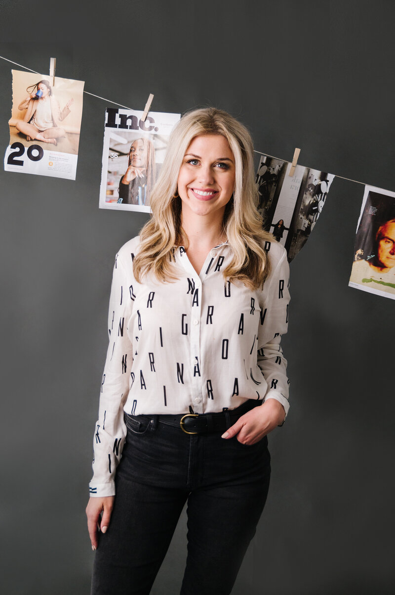 Sarah Klongerbo standing in front of magazine pages hanging on clothespins and wearing black jeans and a white top with letters all over it