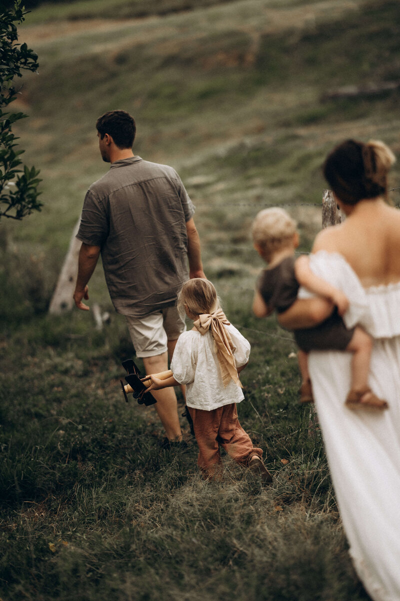 family walking in a green country field the father is leading the way with a little girl holding a toy aeroplane and the mother is holding their baby boy