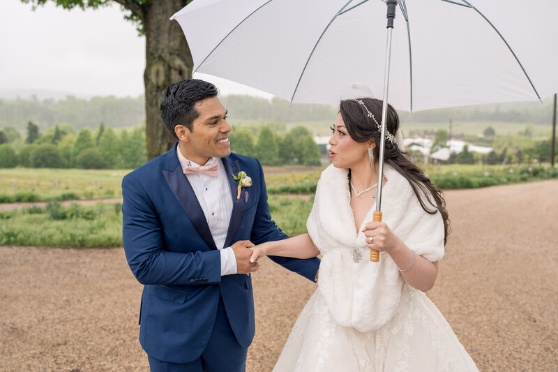 A groom leads his bride in the rain with a white umbrella during their wedding day photos,