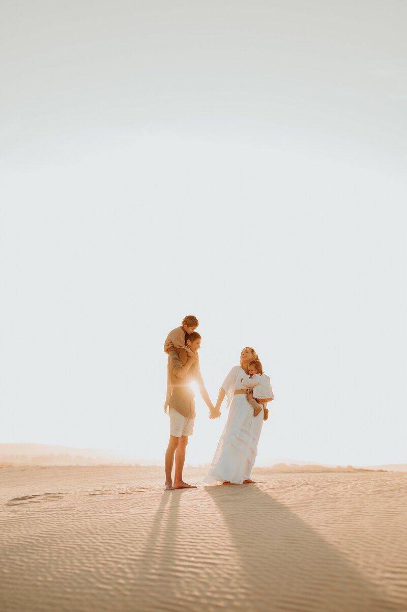 A couple holds hands on the top of a sand dune with the sunsetting behind them. Mom is in a long white dress and is holding one child in her arms. Dad has one child on his shoulders