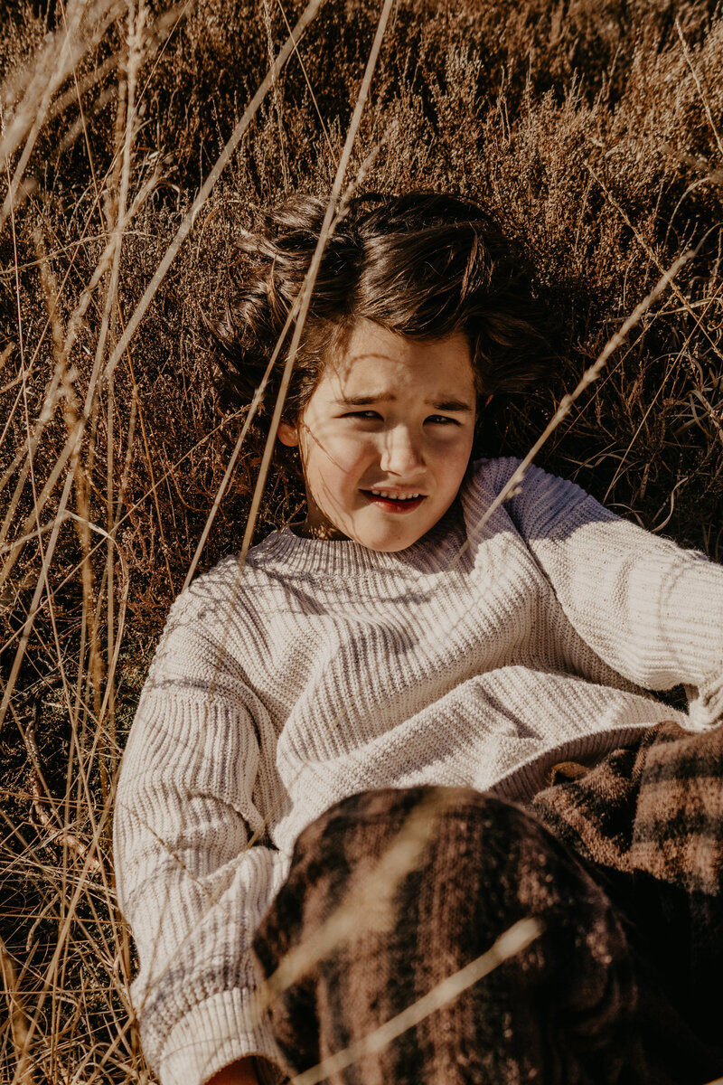 Een jongen ligt ontspannen in het hoge gras, omringd door de natuur en genietend van de rust.