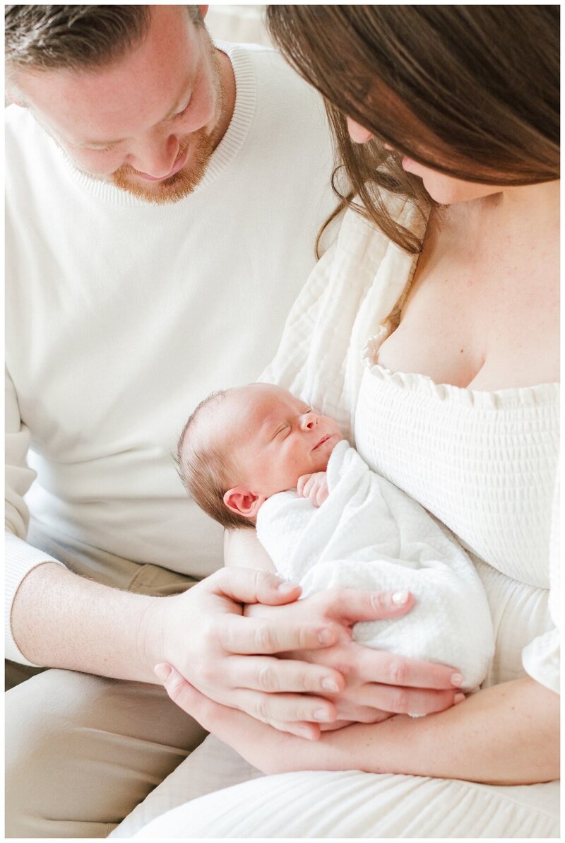A couple snuggles their newborn baby while being photographed by New Jersey Newborn Photographer Kate Voda Photography. The husband is wearing a white sweater and khaki pants, the wife is wearing a cream color dress. The baby is wrapped in a white swaddle.