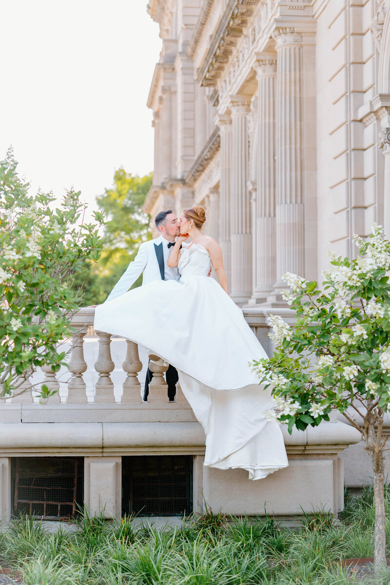 Rhode Island Wedding Photographer | A bride in a flowing white dress sits on a stone railing, lovingly touching noses with a groom in a tuxedo. They are surrounded by lush greenery and a grand, ornate building.