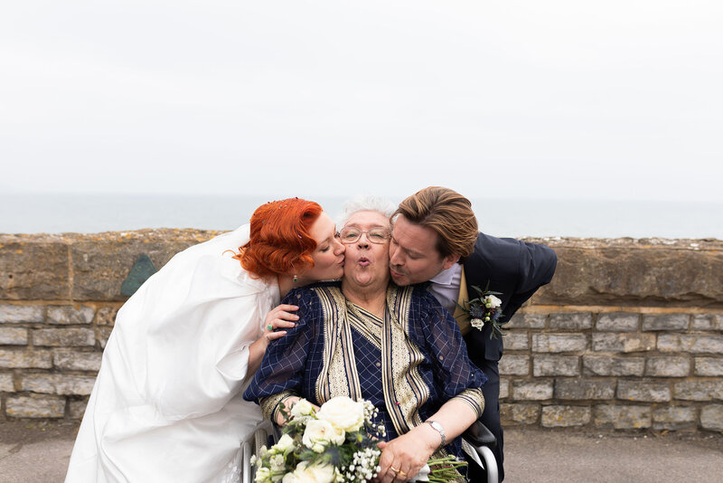 Bride and groom kiss grandma on each cheek. Grandma is in wheel chair making kiss face