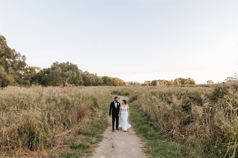 A bride and groom walking through a grassy field with a clear sky.