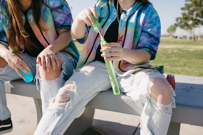 Two friends sitting on a picnic table while one plays with bubbles.