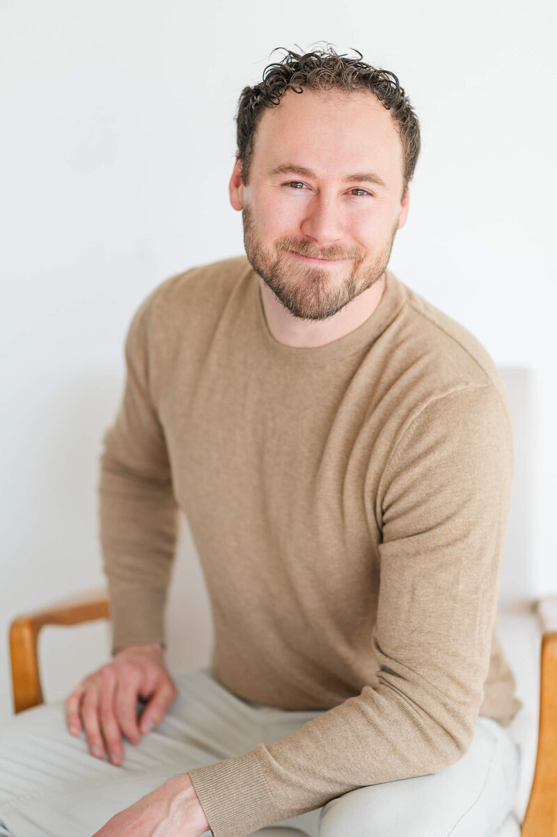 Natural light studio portrait of a man in neutral clothes sitting in a chair smiling at the camera.