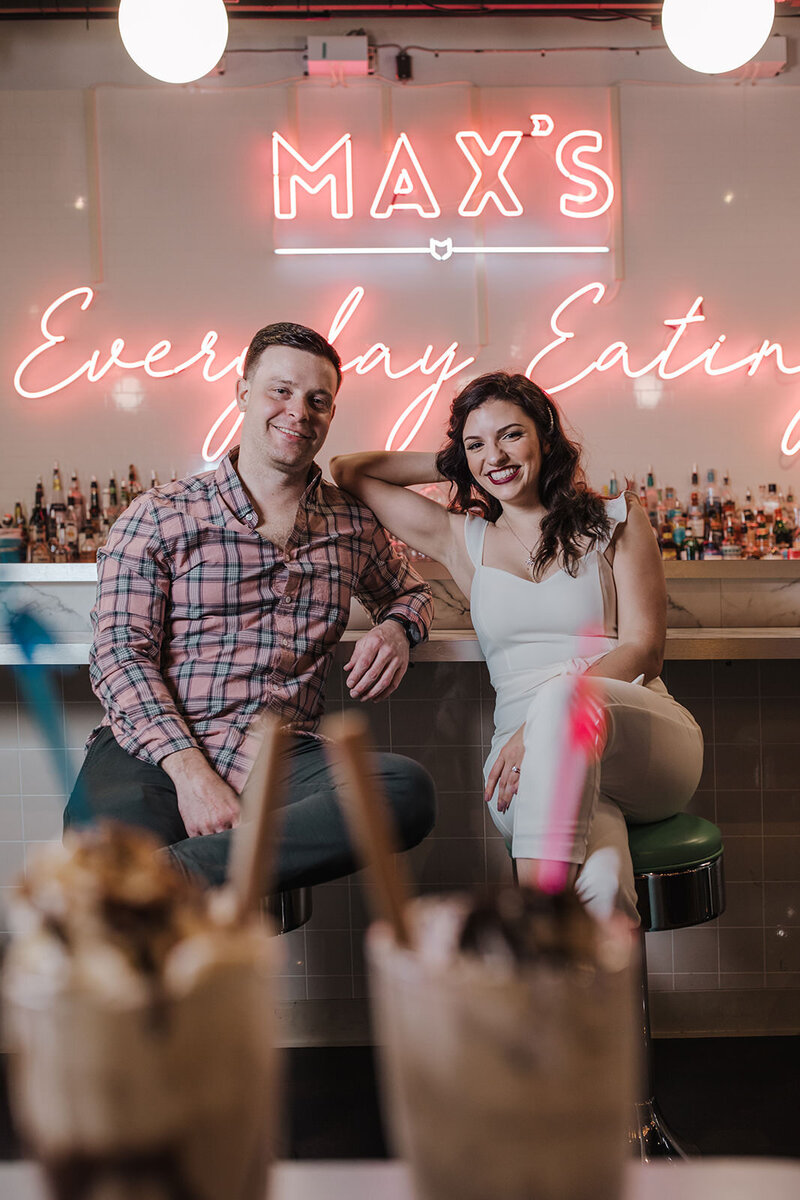 A couple sits indoors, smiling, with milkshakes in the foreground, slightly blurred.