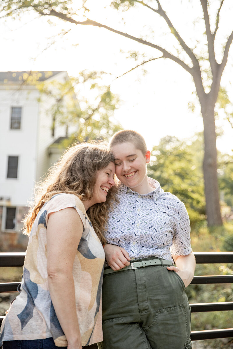 mother and adult child laugh and snuggle together on a bridge during golden hour at sharon mills park in manchester michigan