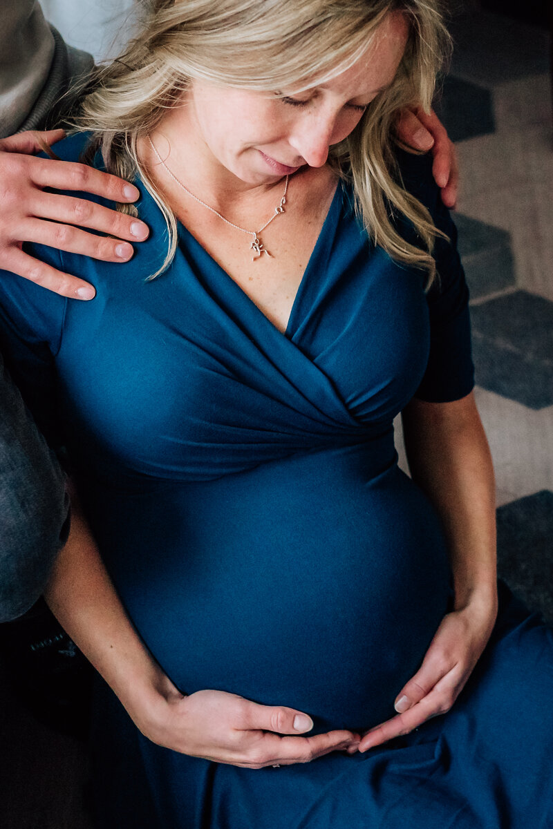 A close-up of a pregnant woman in a deep blue dress, sitting and cradling her belly. Her partner's hands rest gently on her shoulders and stomach, conveying a sense of support and connection.