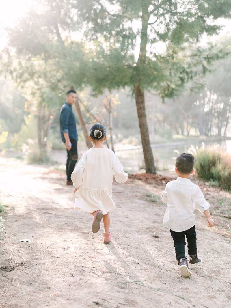 Two children in white clothing run along a sunlit, tree-lined path, exuding joy and energy. A dad in jeans follows, surrounded by a serene, natural setting.