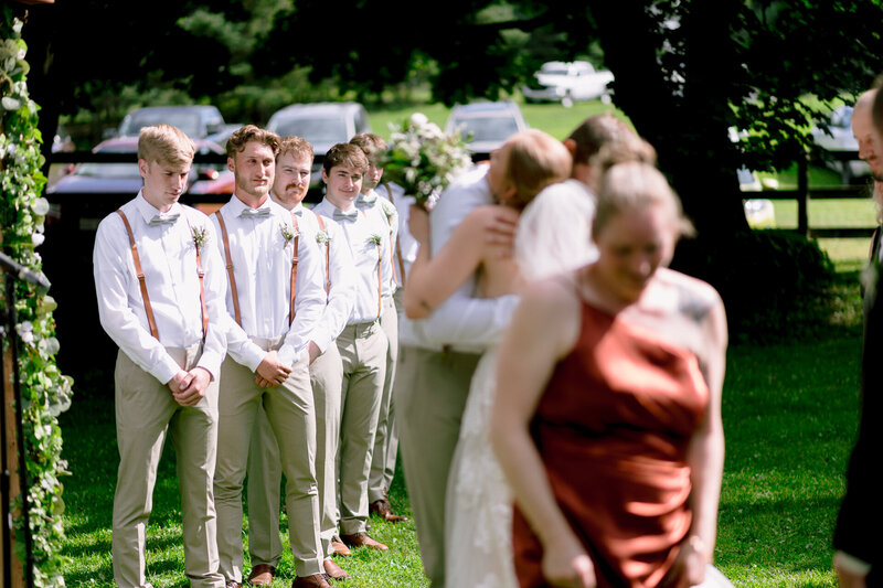 Groomsmen lined up at a wedding ceremony, watching as guests embrace in the foreground.