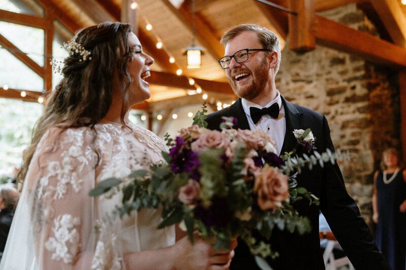 A bride in an elegant gown holds a lush bouquet of roses and greenery, standing beside a groom in a classic tuxedo, amid a warmly lit venue.