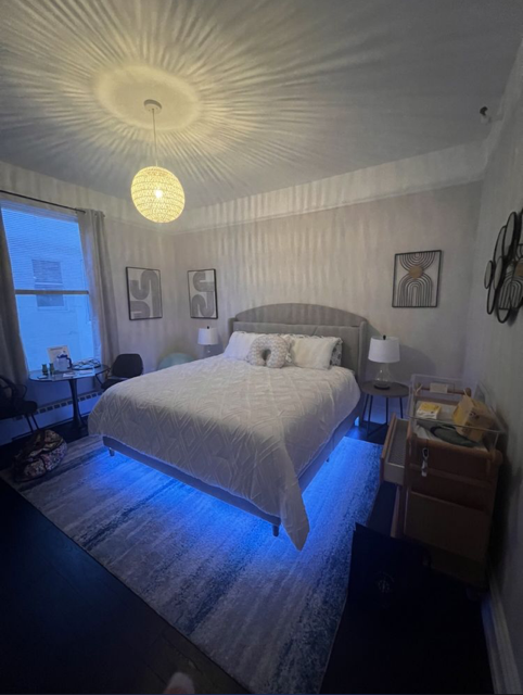 Picture of one of the Village's king studio suites. A room with a king bed piled with pillows with underbed lighting, a hospital-grade bassinet, a table with two chairs next to a window, and abstract art on the walls.