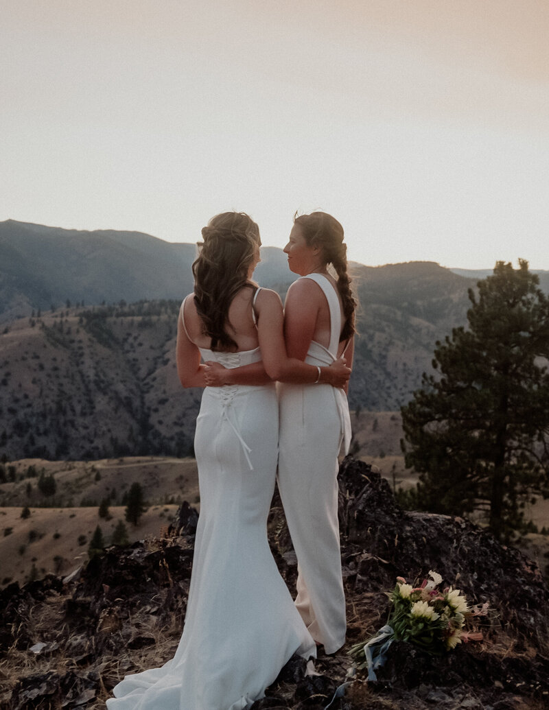 2 brides holding each other on their back looking out in to a mountain view. they both are wearing thin white wedding dress.