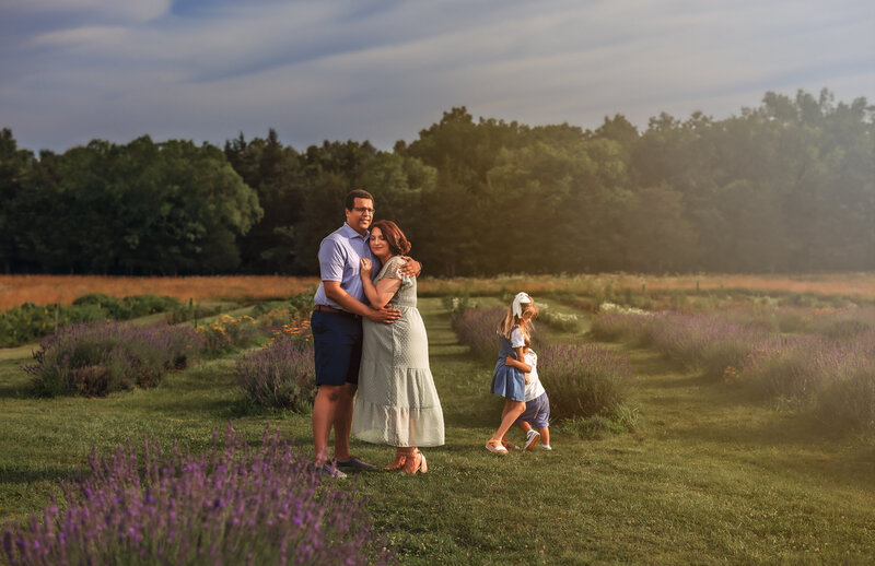 Mom and dad cuddle in close while daughter and son hug in the background of the lavender field in virginia