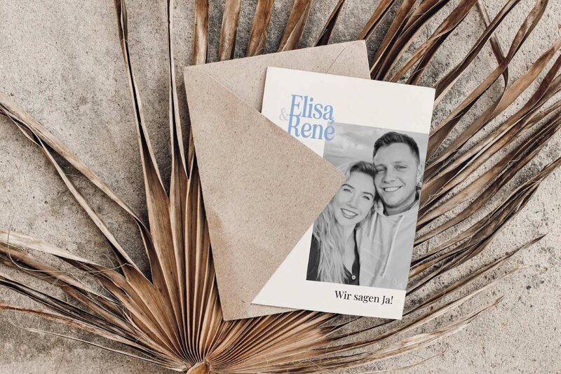 Moderne Hochzeitseinladung mit hellblauer Schrift und schwarz-weiß Foto vom Brautpaar liegt auf einem Palmenwedel
