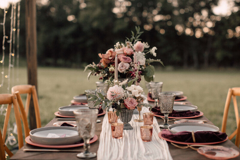 A Hudson Valley outdoor wedding dinner table set with vintage stemware pink and gray plates burgundy velvet napkins and blush and maroon flower arrangements