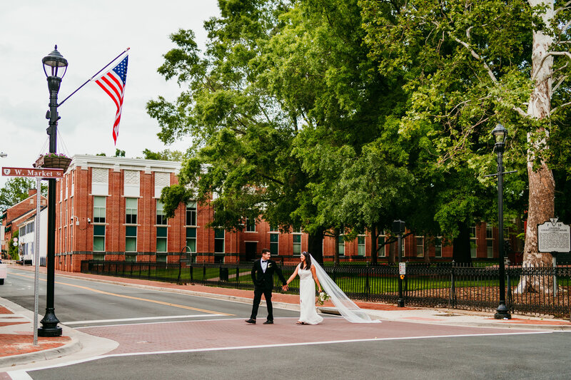 Eloping couple walking hand-in-hand through a crosswalk in front of a courthouse with American flag waving