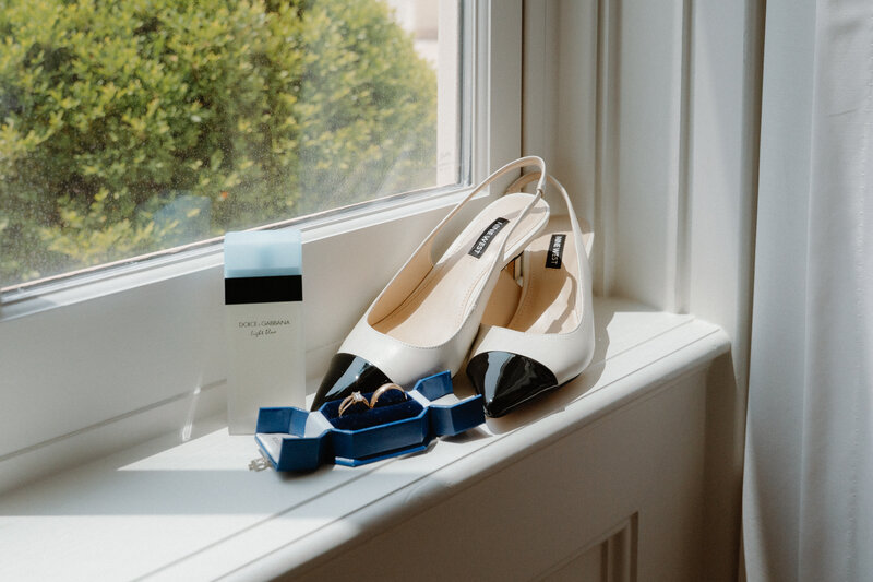 photo of brides shoes, rings, and perfume resting in a window sill with the sun shining through