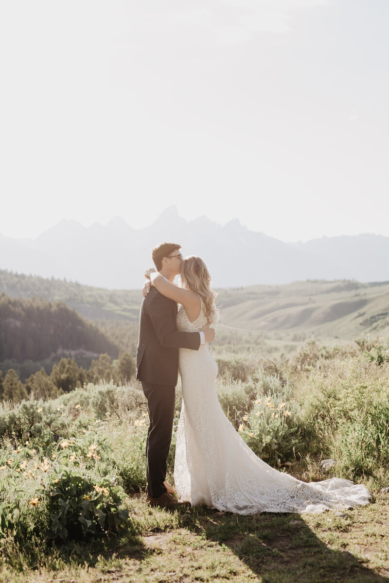 romantic wedding pictures by jackson hole wedding  photographers  with groom holding his brides waist as he leans in and kisses her forehead while the sun begins to set over the grand teton in the distance