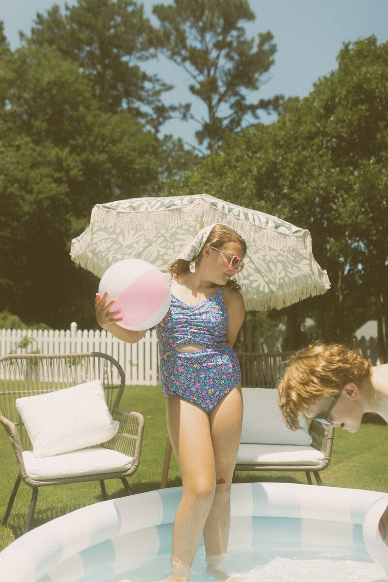 A tween girl holding a beach ball strikes a pose in front of a pool. 