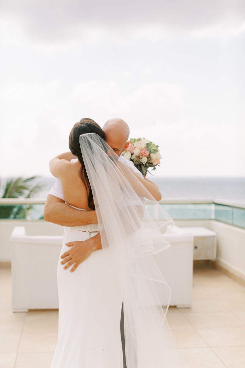 A wedding couple embraces on a tropical rooftop terrace with modern buildings and palm trees in the background.