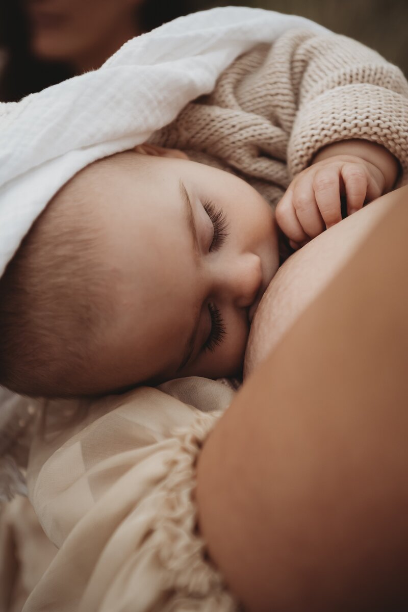 In this serene lifestyle motherhood photography session, an infant with closed eyes is nursing while gently being held, covered with a soft white cloth and wearing a beige knit sweater. Perfectly captured by a Wollongong motherhood photographer, this tender moment embodies the essence of South Coast motherhood portraits.