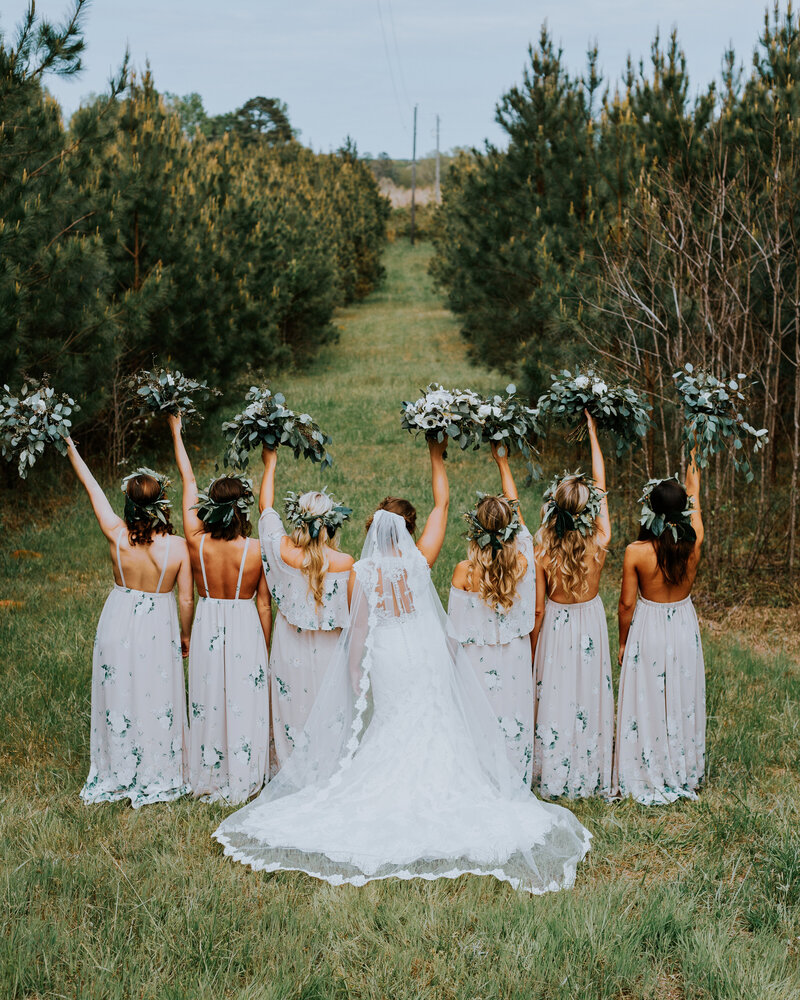 Boho bridesmaids and bride stand in a field with their bouquets in the air, wearing floral crowns at a rustic wedding.